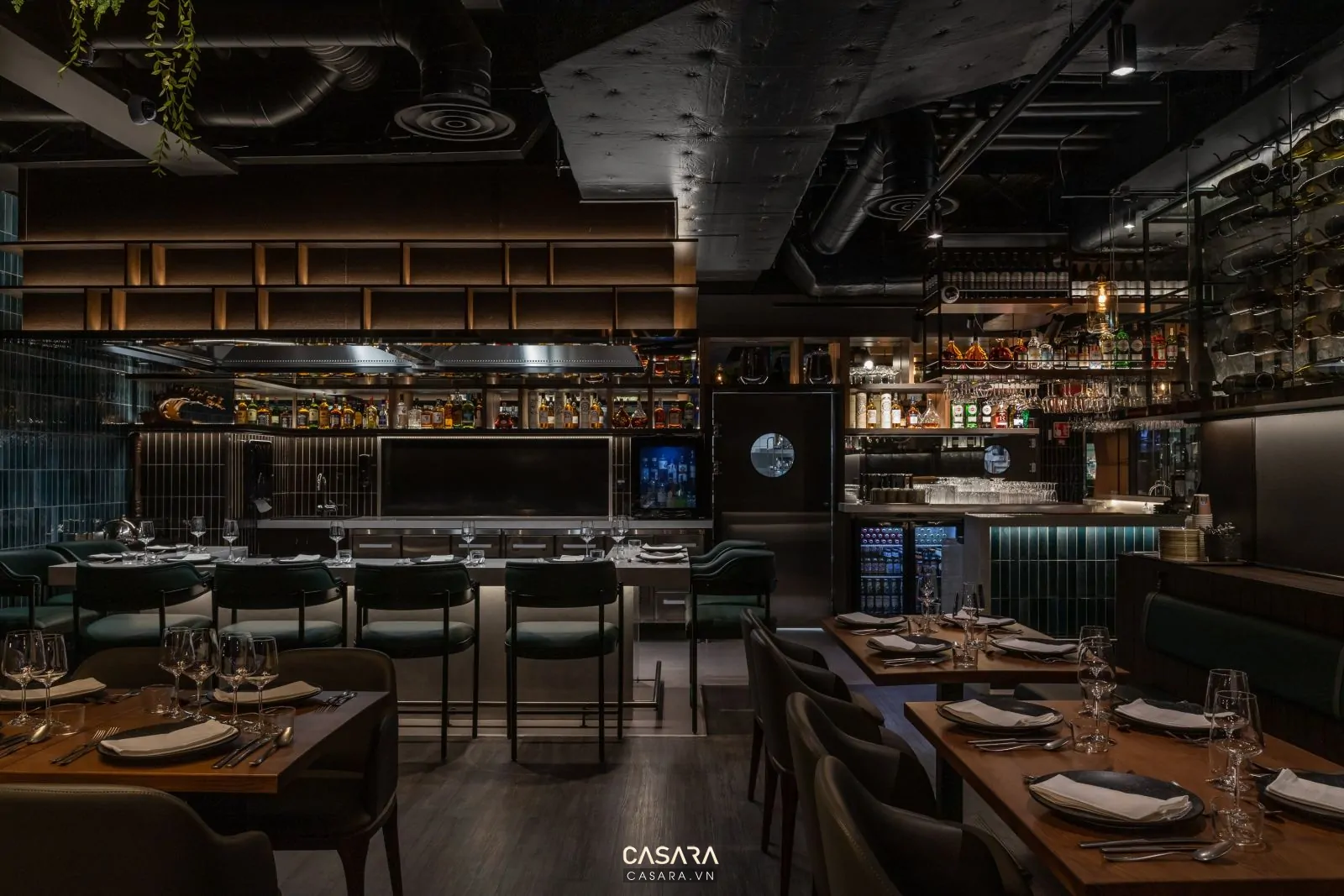 Không gian nhà hàng buổi tối với bàn ăn, đèn thả ánh sáng ấm, vách gỗ hoặc đá, quầy bar và khách dùng bữa