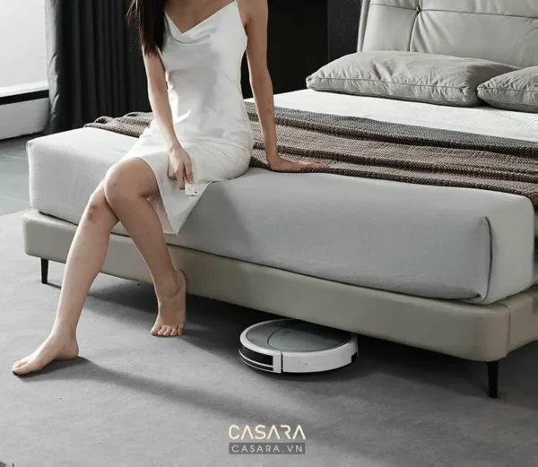 Người phụ nữ thư giãn trên giường, robot hút bụi đang làm việc.