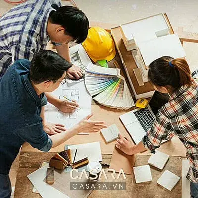Nhóm thiết kế xem đồ án showroom trên bàn họp với bản vẽ mặt bằng, mẫu gỗ đá vải, laptop phối cảnh và ghi chú.