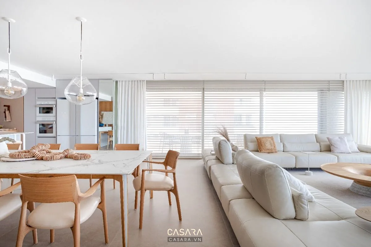Khu khách và bàn ăn mở trong căn hộ sáng màu, có sofa, bàn ăn, bếp liền kề và cửa sổ rộng để tối ưu ánh sáng tự nhiên.