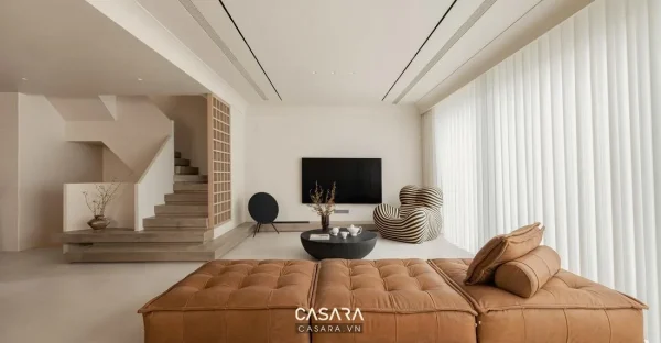 Phòng khách sáng rộng với sofa da nâu, bàn trà tròn, tivi treo tường và rèm trắng lớn