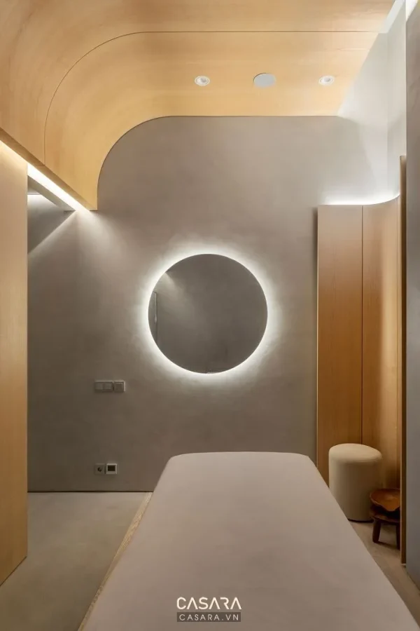 Phòng trị liệu spa có giường massage, gương tròn phát sáng và trần ốp gỗ cong
