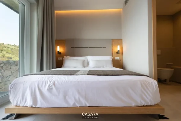 Phòng ngủ nhỏ có giường thấp, vách đầu giường gọn, đèn thả hai bên và cửa kính lấy sáng