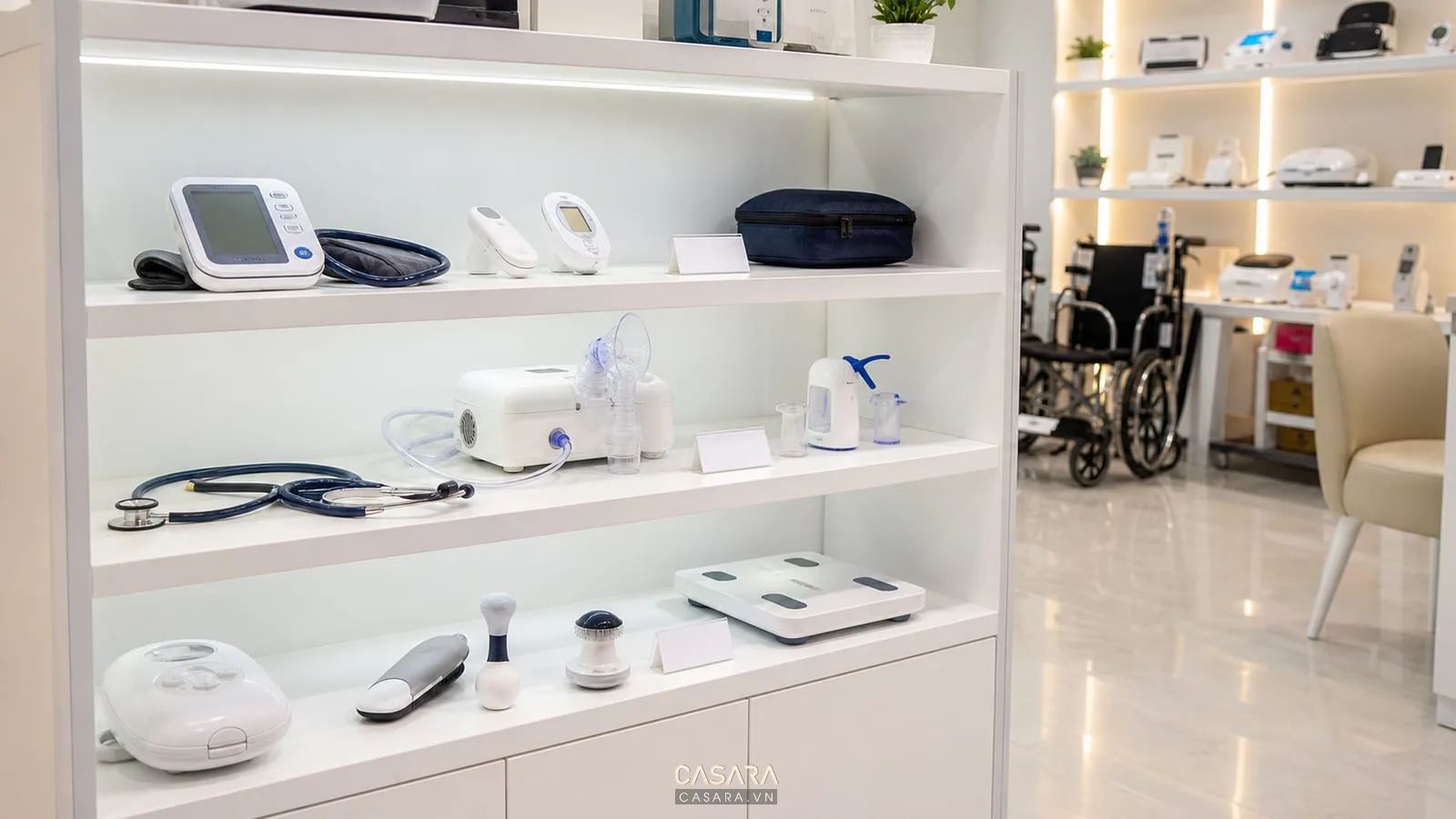 Kệ trưng bày thiết bị y tế nhỏ có máy đo huyết áp, ống nghe, máy xông khí dung, dụng cụ chăm sóc sức khỏe và nhãn thông tin gọn.