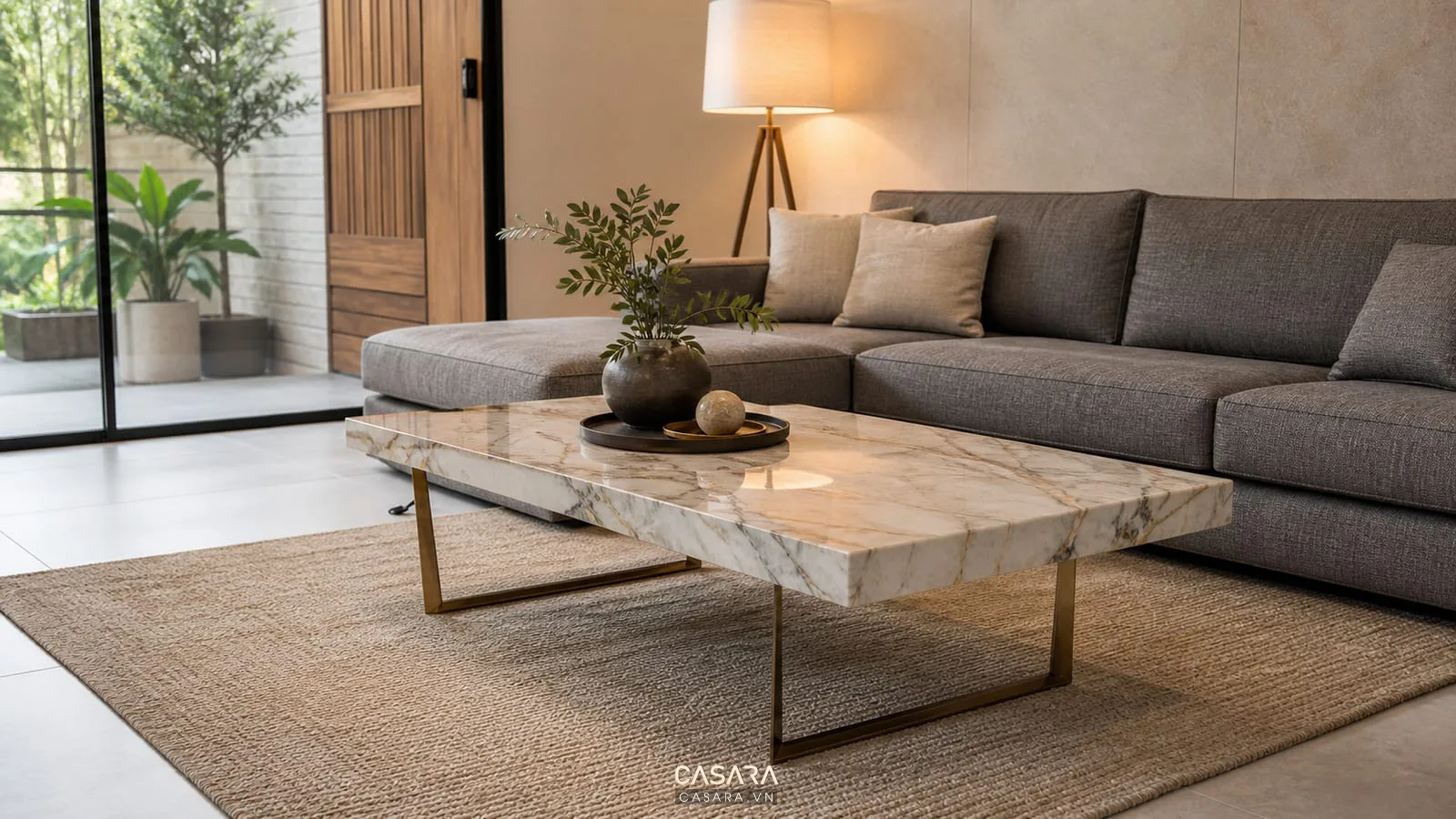 Bàn trà phòng khách bằng đá vân sáng cạnh sofa chữ L màu xám, thảm be, chân kim loại mảnh và đèn sàn ấm