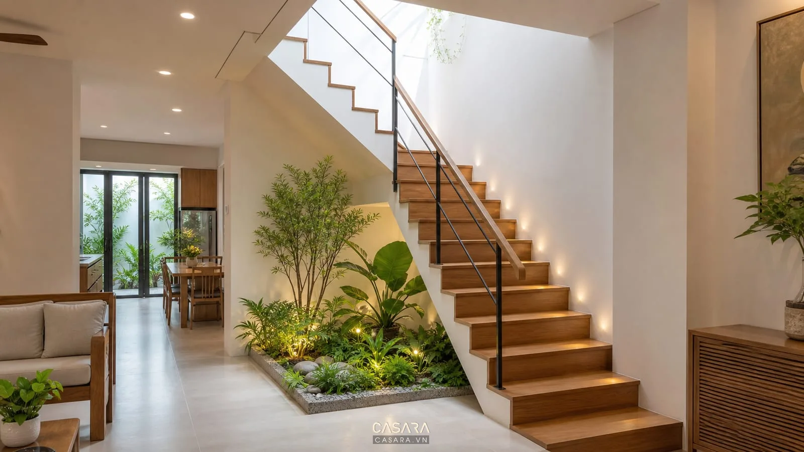 Cầu thang nhà ống có giếng trời, lan can sắt mảnh màu đen, đèn hắt tường, cây xanh nhỏ, bậc gỗ và tường sáng