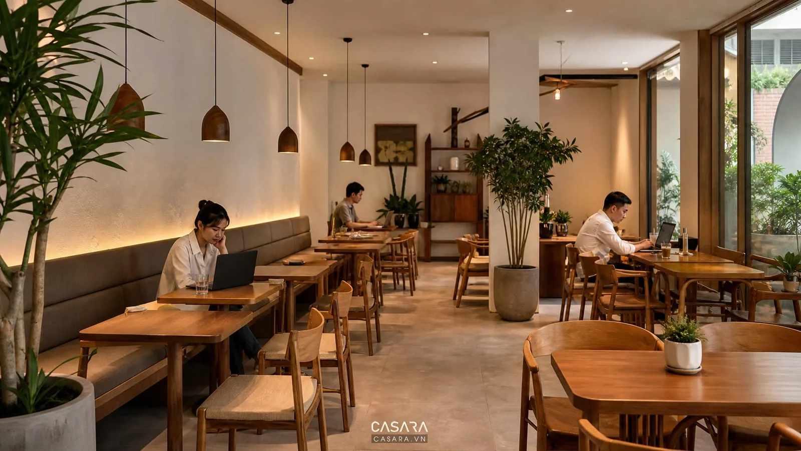 Khu ngồi quán cà phê với bàn ghế cafe gỗ, ghế băng sát tường, đèn thả trần, cây xanh và khách làm việc bằng laptop