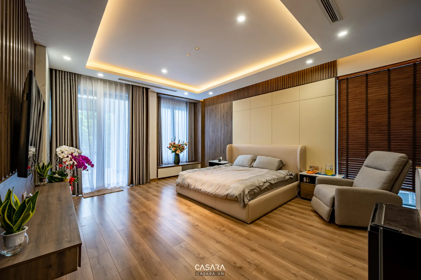 Phòng ngủ hiện đại có giường bọc nệm, sàn gỗ, rèm cửa và trần đèn hắt ấm