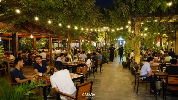 Quán bia sân vườn ngoài trời với bàn ghế gỗ hoặc kim loại, cây xanh, đèn dây treo và khu khách ngồi uống bia buổi tối