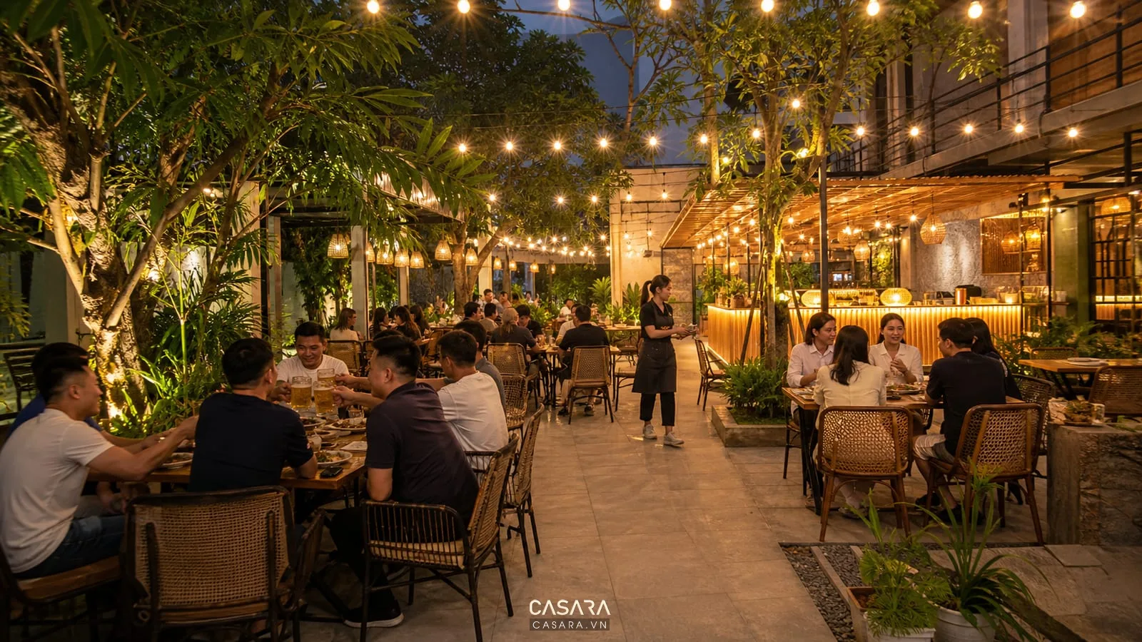 Quán bia sân vườn buổi tối với đèn dây treo, quầy bia sáng ấm, bàn ghế ngoài trời, cây xanh và khách ngồi uống bia
