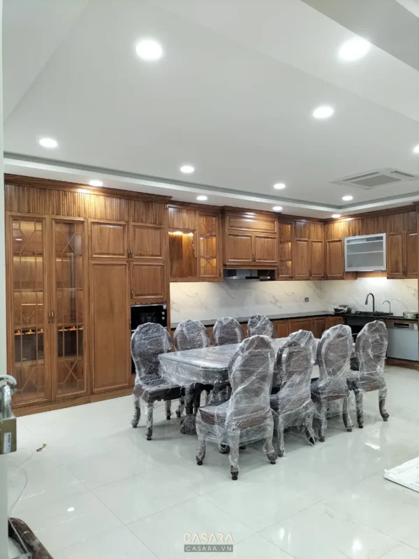 Phòng bếp gỗ với hệ tủ chữ L, bàn ăn và ghế còn bọc nilon sau thi công
