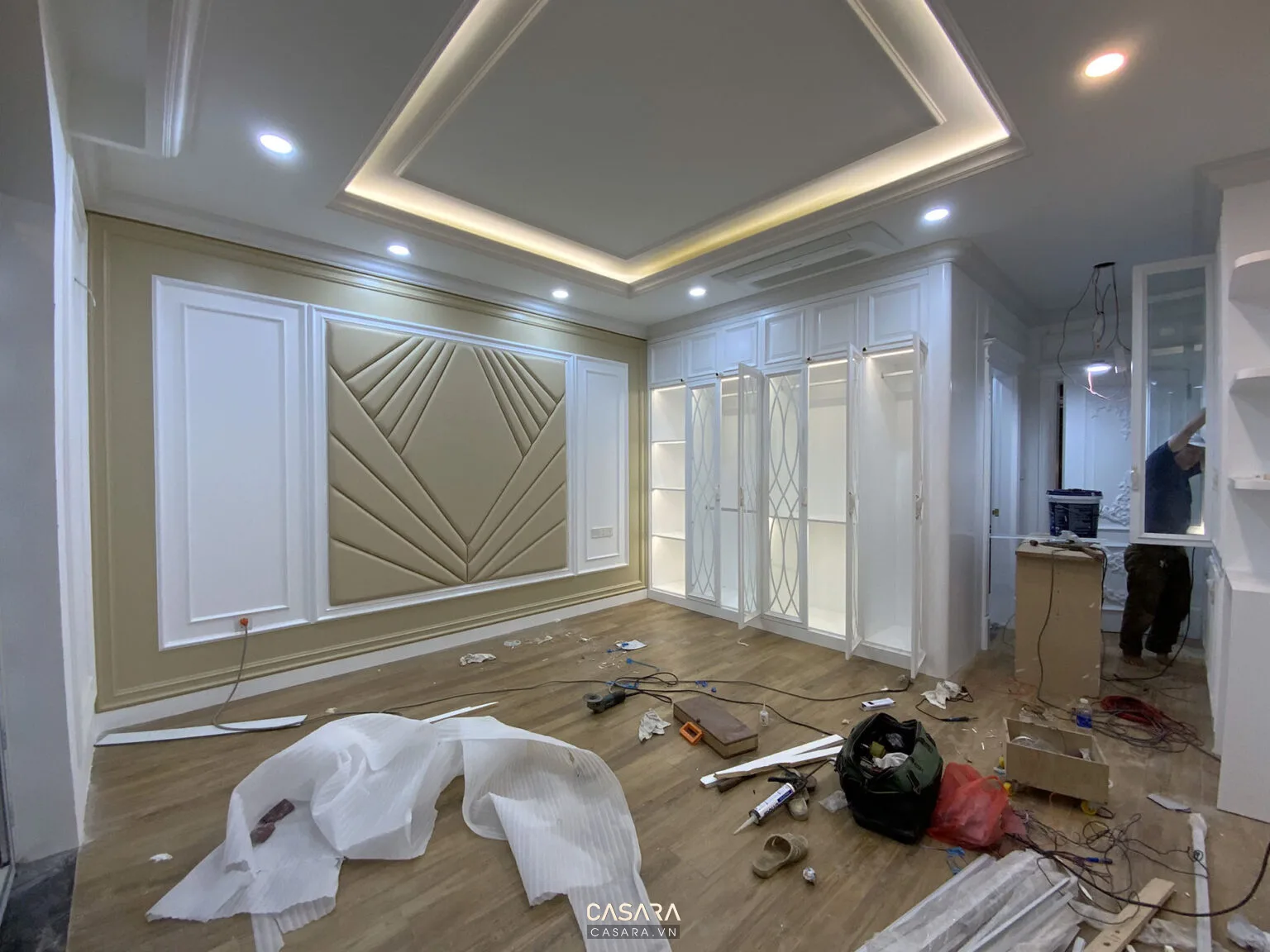Phòng ngủ đang thi công với tủ áo trắng, vách đầu giường bọc nệm và trần giật cấp có đèn hắt