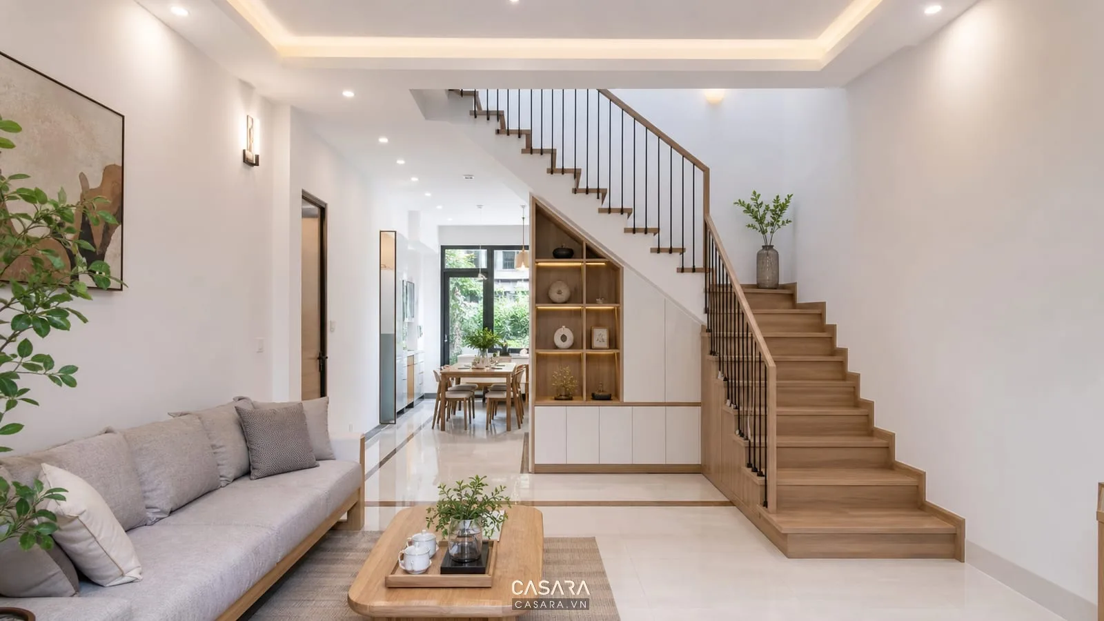 Cầu thang cho nhà ống 4m nhìn từ cửa chính với phòng khách sáng, lan can kính, tủ gầm thang gọn, bếp phía sau và nội thất hiện đại