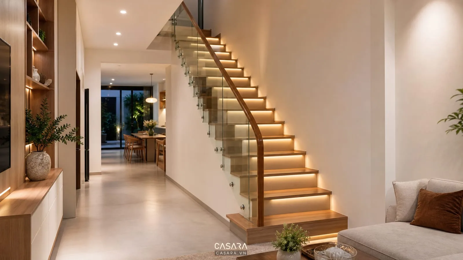 Cầu thang nhà ống 2 tầng có đèn hắt bậc, tay vịn gỗ, lan can kính, tường sáng và không gian buổi tối ấm áp