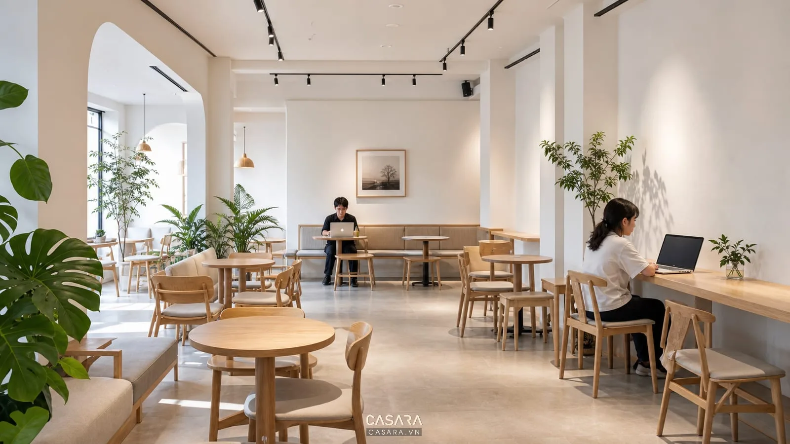 Quán coffee tối giản có bàn ghế cafe gỗ sáng tường trắng đèn thanh ray cây xanh và khách dùng laptop