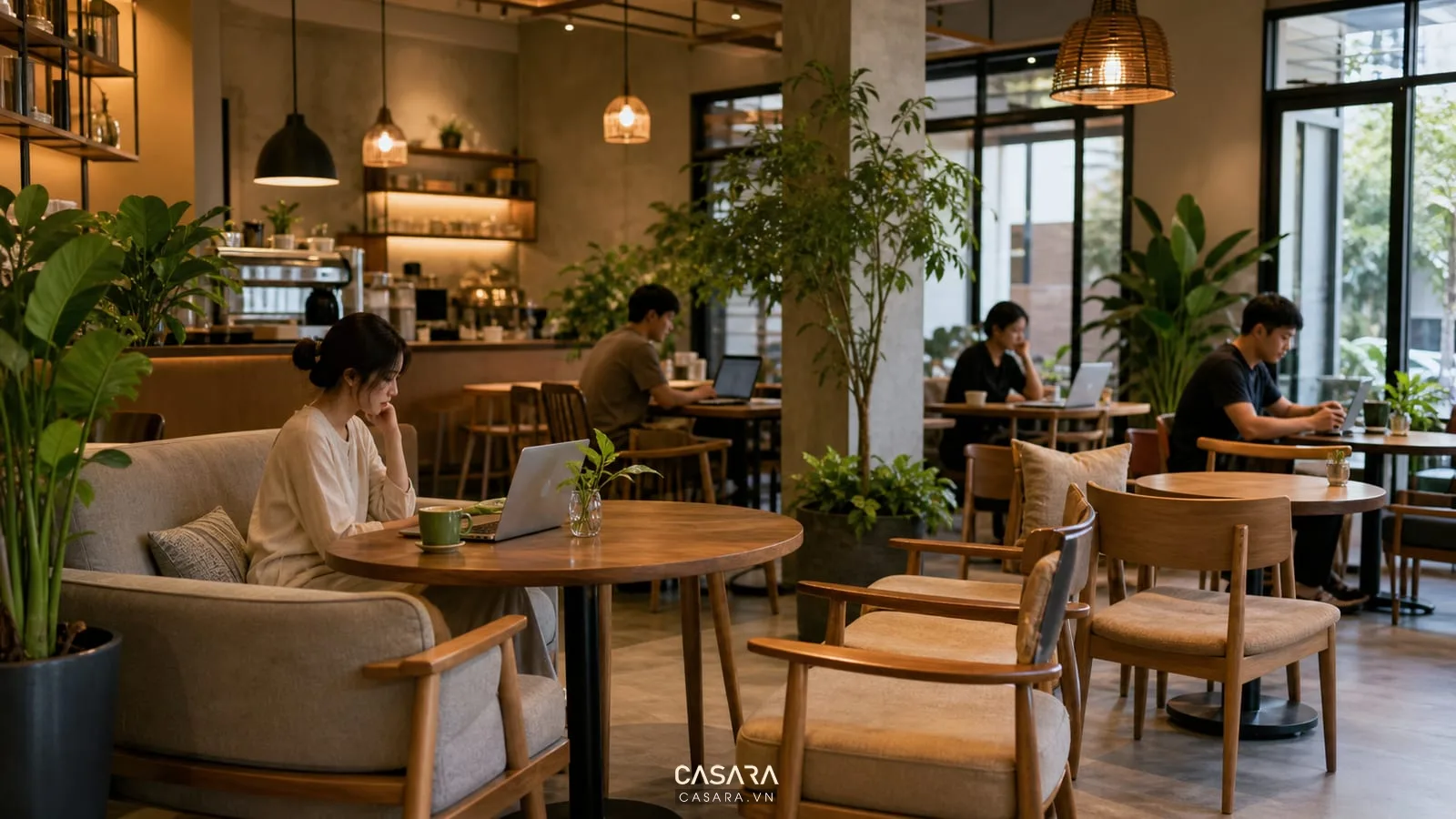 Bàn ghế quán cà phê gồm sofa nhỏ ghế gỗ bàn tròn khách dùng laptop cây xanh và đèn thả ấm