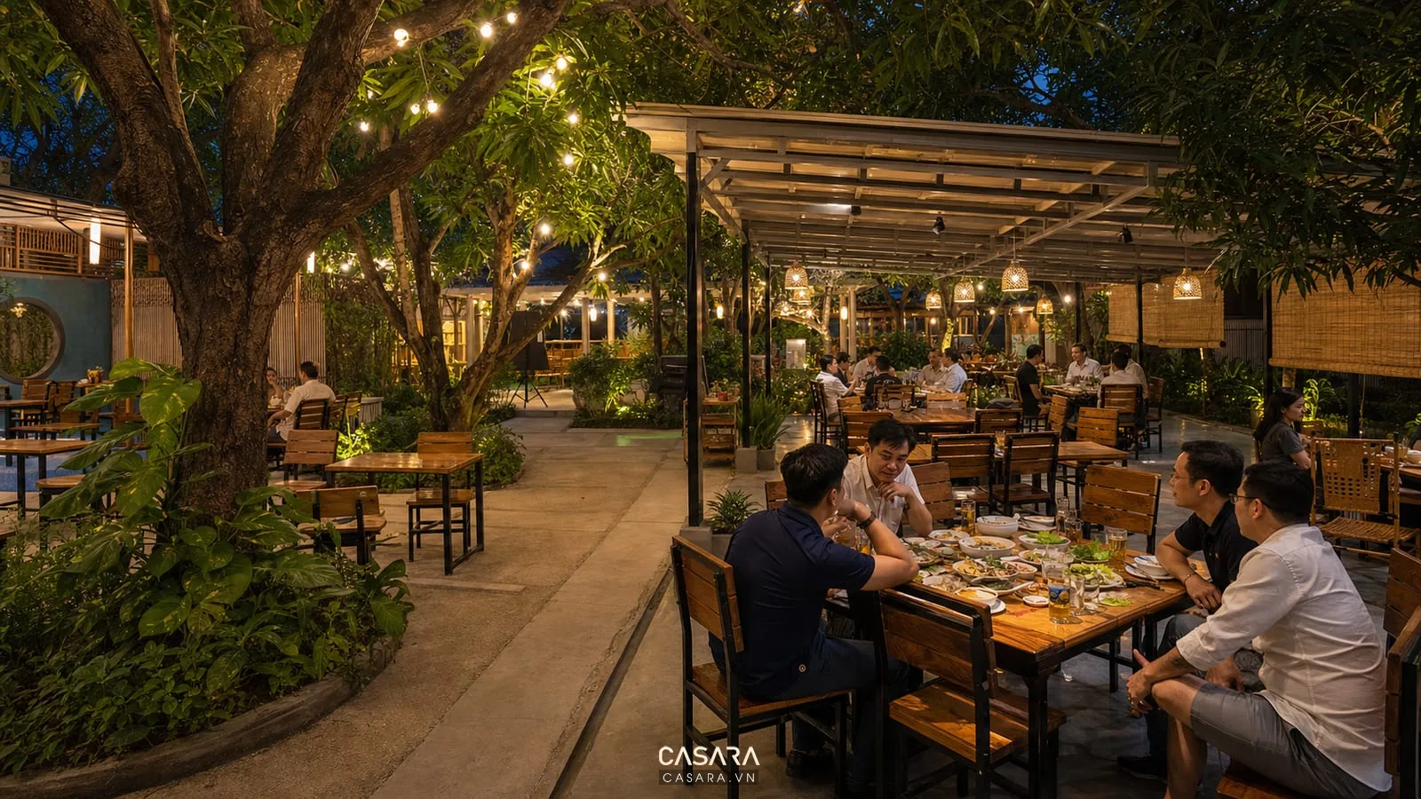 Quán nhậu độc lạ sân vườn có bàn gỗ ngoài trời, cây xanh lớn, đèn dây ánh sáng vàng, mái che nhẹ và nhóm khách ăn tối