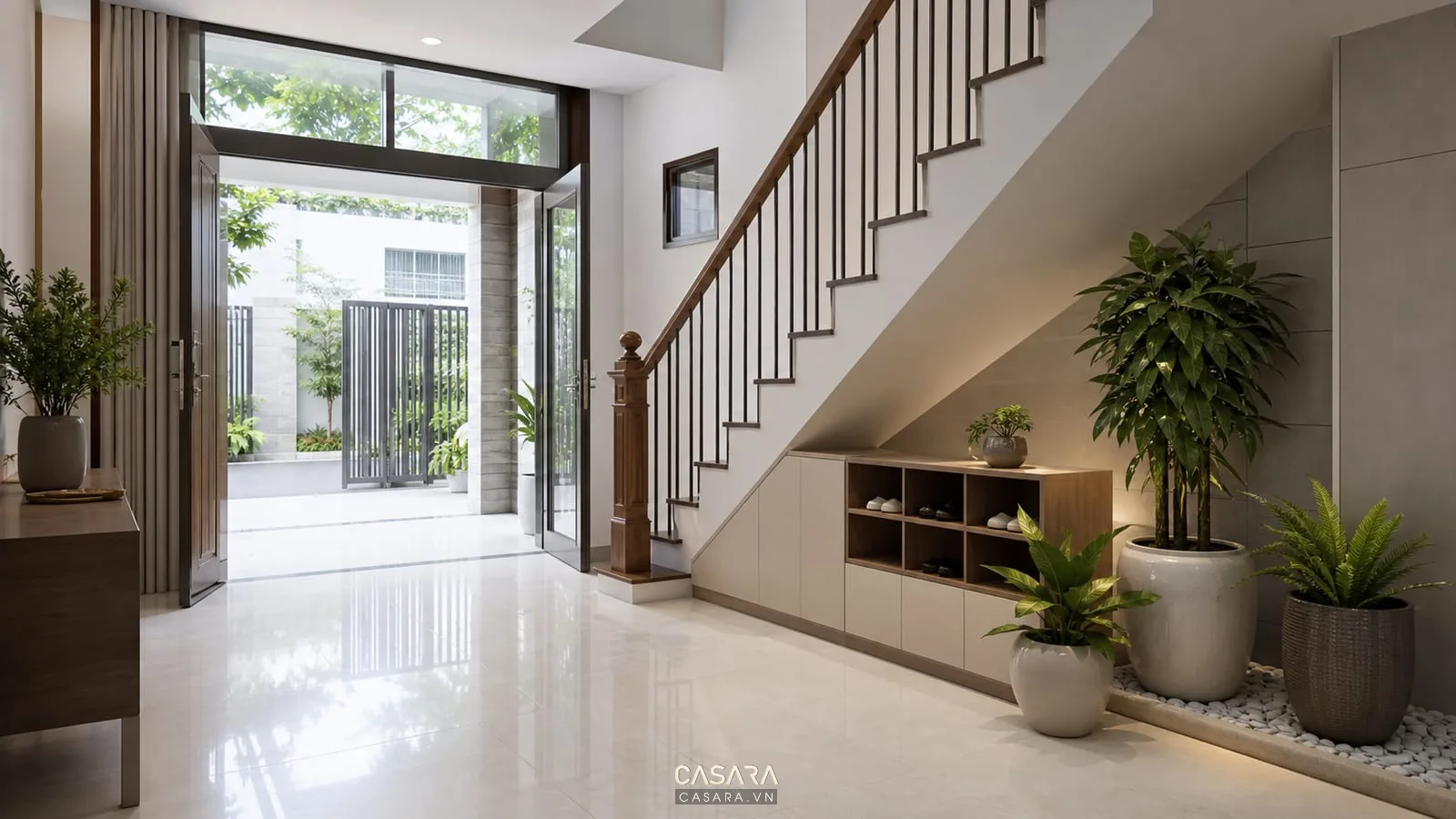 Trang trí cây xanh gầm cầu thang gần cửa vào với chậu cây xanh dưới cầu thang, tủ giày thấp, sàn gạch sáng và nhà phố hiện đại