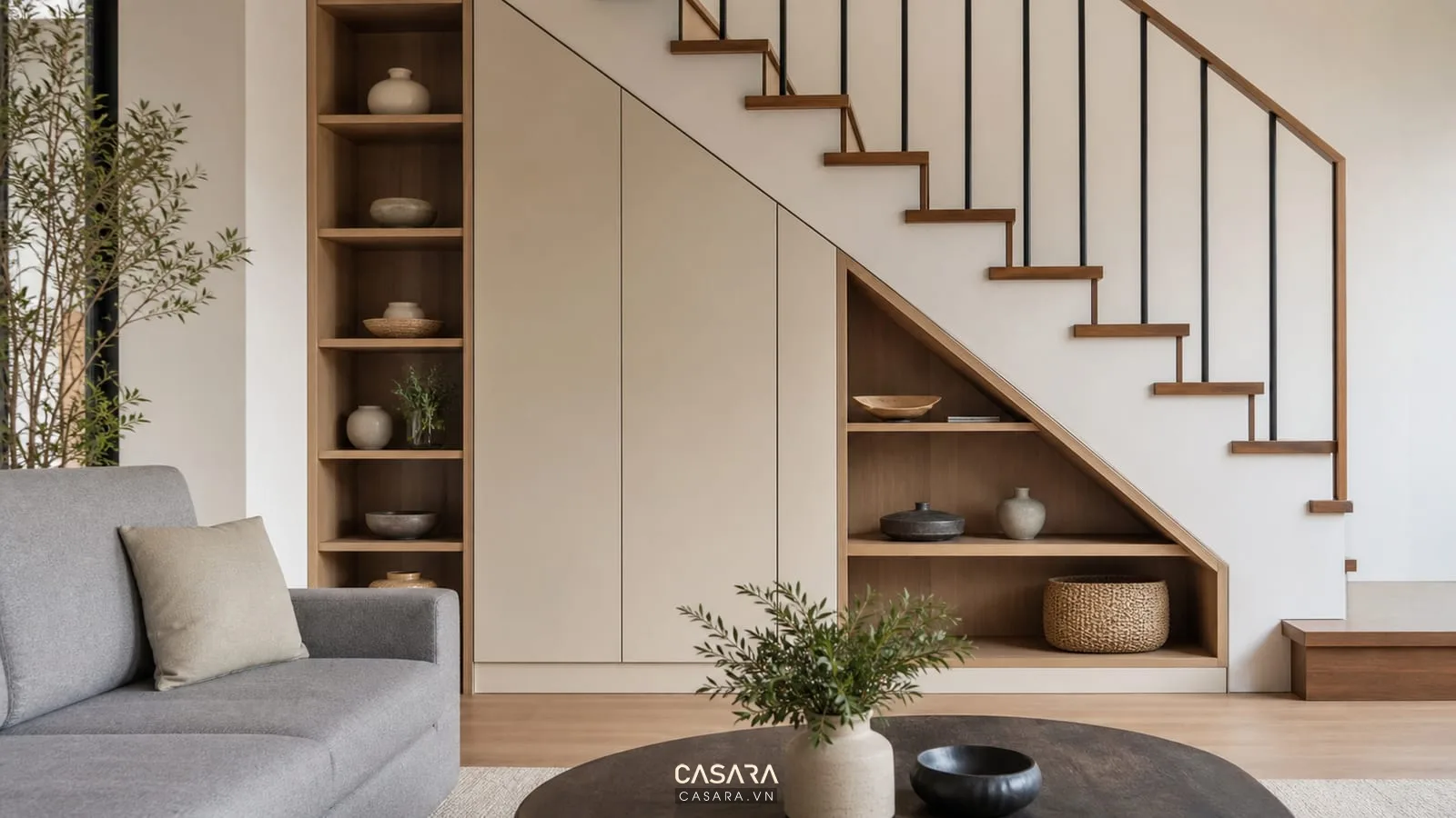 Trang trí gầm cầu thang phòng khách bằng tủ âm màu sáng, kệ gỗ mở, sofa vải xám, bàn trà thấp và cầu thang bậc gỗ