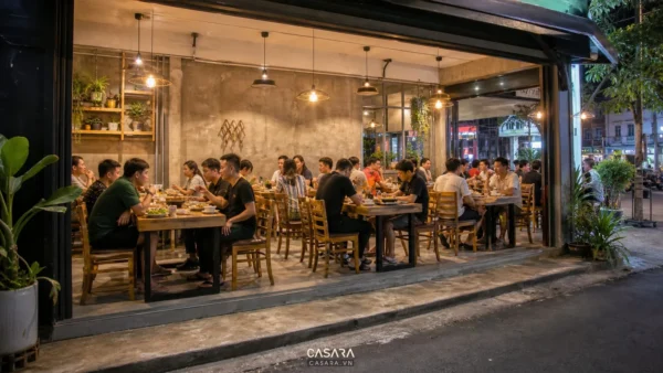 Quán nhậu đơn giản ngoài phố với bàn ghế gỗ mộc, đèn thả ánh vàng, tường xi măng và khu ăn uống đông khách