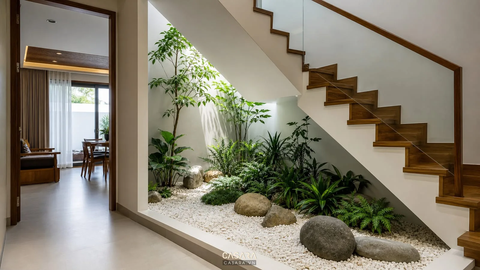 Gầm cầu thang nhà ống có tiểu cảnh khô với sỏi trắng, cây xanh chịu bóng, tường sơn trung tính, cầu thang bậc gỗ và ánh sáng giếng trời